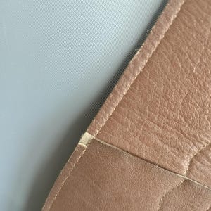 May include: Close-up of a section of brown leather, possibly from a bag or upholstery. The leather has a textured surface with visible stitching along the edges. A light gray fabric is visible in the background.