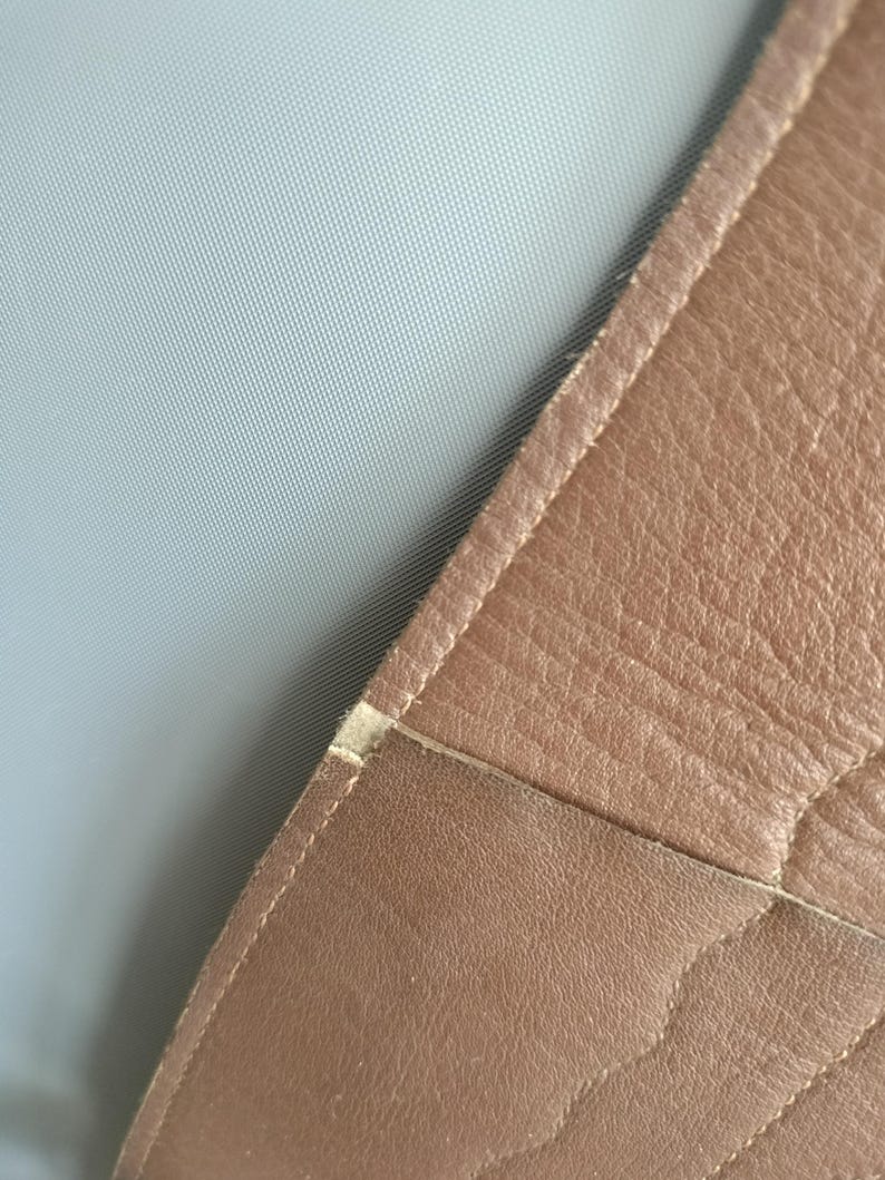 May include: Close-up of a section of brown leather, possibly from a bag or upholstery. The leather has a textured surface with visible stitching along the edges. A light gray fabric is visible in the background.