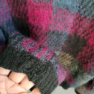 May include: Close-up of a sweater with a plaid pattern in shades of black, teal, and magenta. The cuff is black with teal and magenta accents. The sweater has a soft, textured appearance.