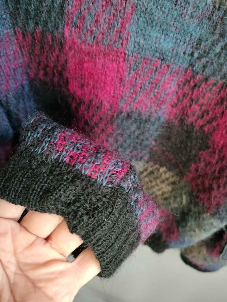 May include: Close-up of a sweater with a plaid pattern in shades of black, teal, and magenta. The cuff is black with teal and magenta accents. The sweater has a soft, textured appearance.