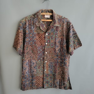 Peut inclure: Une chemise à manches courtes boutonnée avec un motif patchwork dans des tons de marron, vert et bleu. La chemise a un col chemise et est suspendue à un cintre en bois sur un fond gris. Le tissu a un léger éclat.