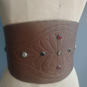 May include: A wide, brown leather belt with a decorative floral pattern and studded accents. The belt features a series of red, black, and silver-toned studs. The belt is displayed on a mannequin.