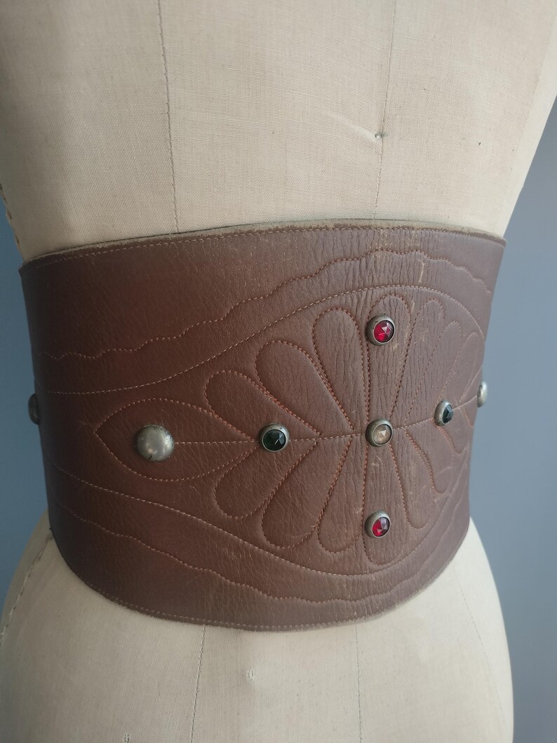 May include: A wide, brown leather belt with a decorative floral pattern and studded accents. The belt features a series of red, black, and silver-toned studs. The belt is displayed on a mannequin.