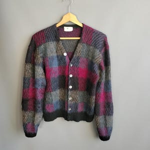 May include: A multi-colored, button-down cardigan sweater with a checkerboard pattern in shades of gray, blue, and magenta. The sweater is hanging on a wooden hanger against a gray background. The sweater has a V-neck and long sleeves.