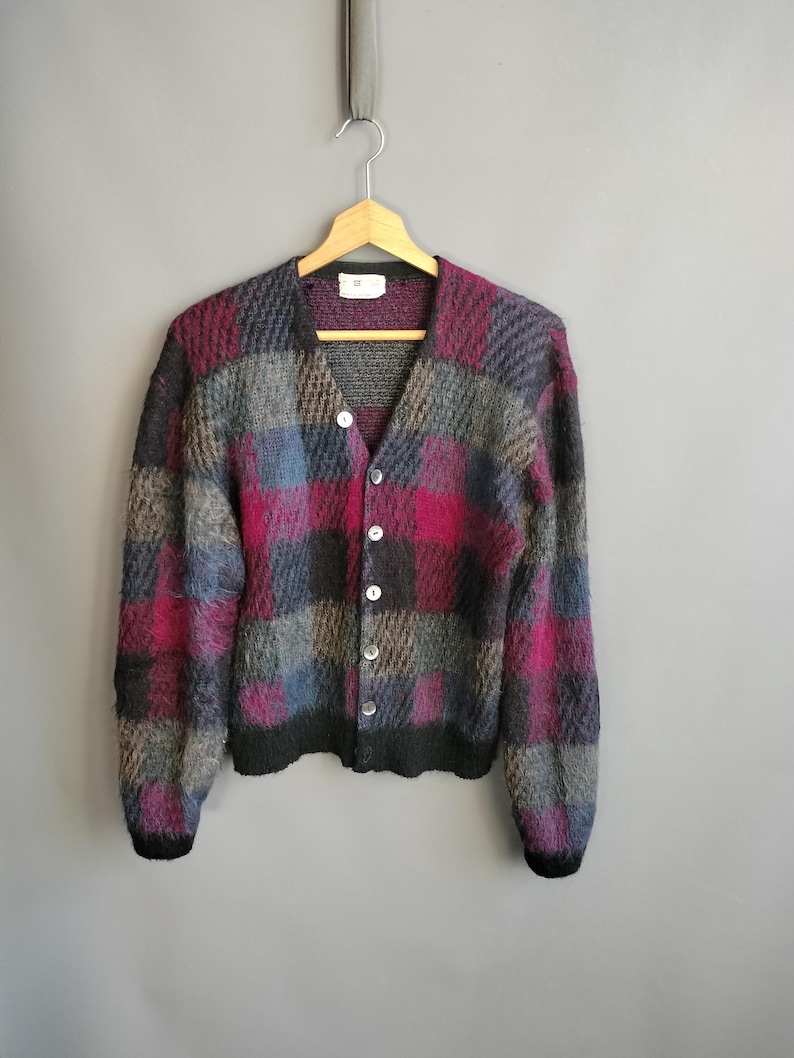 May include: A multi-colored, button-down cardigan sweater with a checkerboard pattern in shades of gray, blue, and magenta. The sweater is hanging on a wooden hanger against a gray background. The sweater has a V-neck and long sleeves.