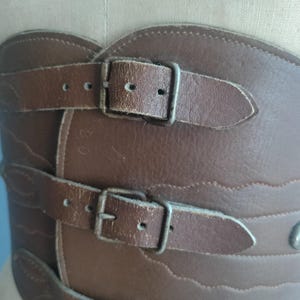 May include: Brown leather corset with three buckled straps. The corset has a decorative wavy edge and metal hardware. The leather appears worn, giving it a vintage aesthetic. The corset is designed to cinch the waist.