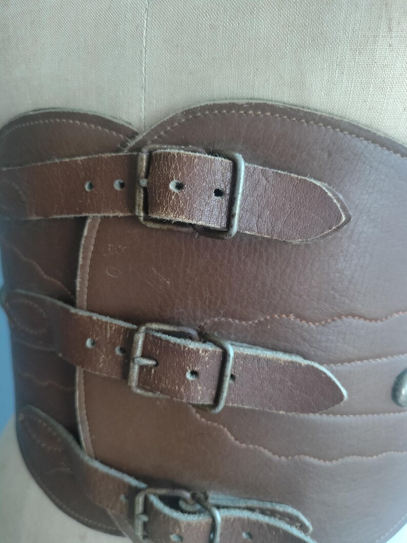 May include: Brown leather corset with three buckled straps. The corset has a decorative wavy edge and metal hardware. The leather appears worn, giving it a vintage aesthetic. The corset is designed to cinch the waist.