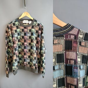 May include: A vintage crew neck sweater with a geometric pattern in shades of black, brown, pink, green, and blue. The sweater has a ribbed collar, cuffs, and hem. The brand name "DANIELAPNES" is visible on the collar.
