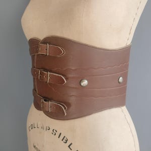 May include: A brown leather corset belt with three buckle closures and decorative stitching. The wide belt features two silver rivets and is displayed on a mannequin. The belt is a vintage style.