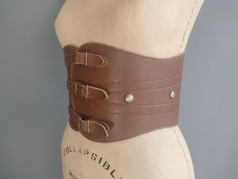 May include: A brown leather corset belt with three buckle closures and decorative stitching. The wide belt features two silver rivets and is displayed on a mannequin. The belt is a vintage style.