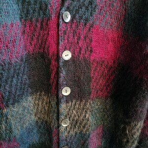 May include: A close-up of a button-down cardigan with a plaid pattern. The sweater features a mix of colors, including teal, burgundy, black, and tan. The buttons are a pearlescent color. The fabric appears to be a soft, fuzzy material.