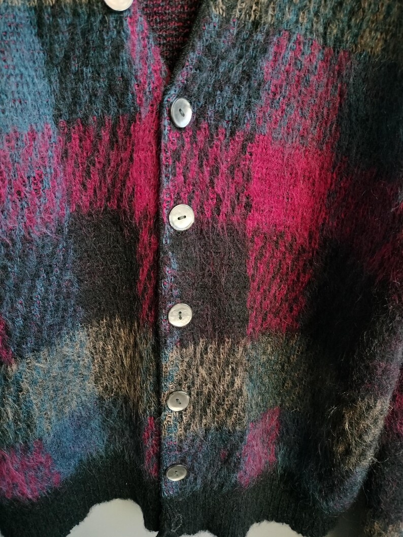 May include: A close-up of a button-down cardigan with a plaid pattern. The sweater features a mix of colors, including teal, burgundy, black, and tan. The buttons are a pearlescent color. The fabric appears to be a soft, fuzzy material.