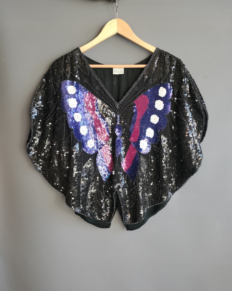 May include: A black sequined top featuring a large butterfly design in shades of purple, pink, and white. The butterfly is positioned centrally on the front of the top.