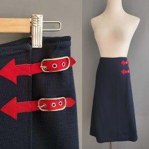 May include: A navy blue skirt featuring red arrow-shaped details and matching red belt-style fastenings. The skirt is displayed on a mannequin and a hanger, highlighting the design and fit. The skirt has a classic A-line shape.