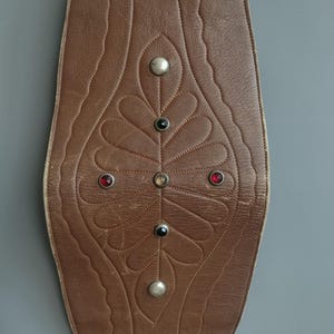May include: Brown leather belt with decorative leaf pattern and silver-colored studs. The belt features a central design with red, black, and clear gemstones. The edges have a scalloped design.