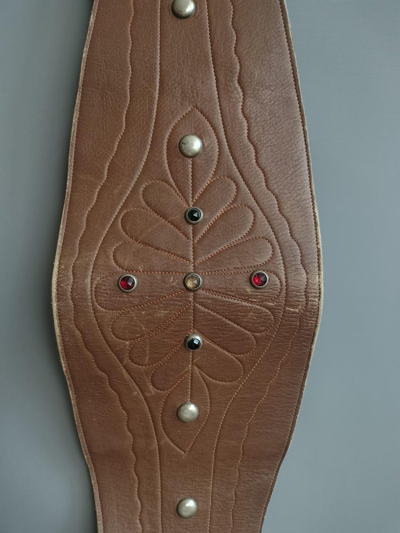 May include: Brown leather belt with decorative leaf pattern and silver-colored studs. The belt features a central design with red, black, and clear gemstones. The edges have a scalloped design.