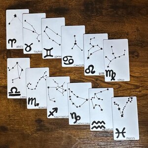 Puede incluir: Un conjunto de tarjetas rectangulares blancas, cada una con un diseño de constelación negra y un símbolo del zodíaco. Las tarjetas están dispuestas sobre una superficie de madera marrón, con los signos del zodíaco Aries, Tauro, Géminis, Cáncer, Leo, Virgo, Libra, Escorpio, Sagitario, Capricornio, Acuario y Piscis.