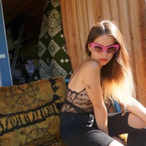 Può includere: Una donna indossa occhiali da sole rosa a occhi di gatto e un top di pizzo nero. Gli occhiali da sole hanno una tinta marrone chiaro. Indossa jeans neri con ginocchia strappate. Lo sfondo include un muro di legno e tessuto fantasia.