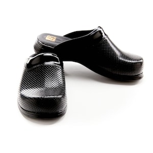 May include: A pair of black slip-on clogs featuring a perforated texture across the upper surface. The shoes have a rounded toe, a thick sole, and a gold logo printed on the inner heel area. The design is simple, functional, and solid black.