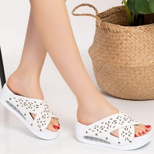 May include: White platform sandals with a cut-out design. The sandals have a criss-cross strap and are embellished with small silver studs. The image also shows a decorative plant in a woven basket.