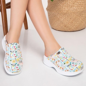 May include: White clogs with a medical-themed pattern featuring cartoon doctors, medical tools, and text. The clogs have a white sole and a slip-on design. The background includes a plant in a woven basket.