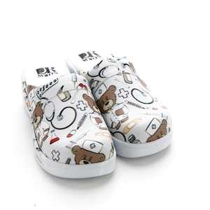 Puede incluir: Zuecos blancos con temática médica. Los zuecos presentan un patrón repetido de osos de peluche marrones, estetoscopios, jeringas, vendas y cruces. Los zuecos tienen una suela blanca y el texto "DR MOON" en la parte superior.