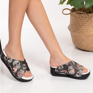 Peut inclure: Une paire de sandales à enfiler noires avec des brides croisées ornées d'un motif de crânes et de fleurs en blanc, gris et rouge. Ces sandales présentent une épaisse semelle plateforme noire avec une couche intermédiaire blanche et un bout ouvert.