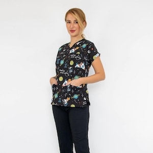 May include: Black scrub top and pants set. The top features a V-neck and pockets, adorned with a colorful space-themed print of rockets, planets, and stars. The matching pants are solid black. The outfit is paired with black clogs.