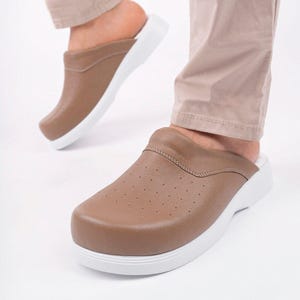 Puede incluir: Un par de zuecos sin cordones de color topo con una parte superior texturizada, pequeños orificios de ventilación en la parte superior y una suela blanca gruesa y sólida. El calzado tiene una puntera redondeada y el talón abierto y funcional.
