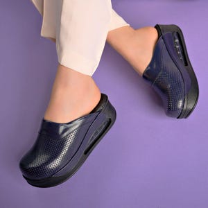 May include: Navy blue perforated clogs with a black sole and a visible air cushion. The clogs have a glossy finish and are worn with cream-colored pants. The background is a solid purple.