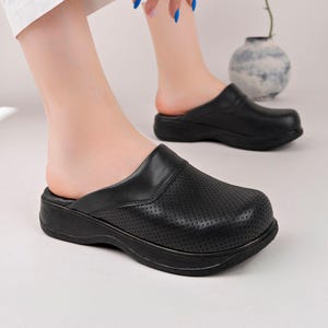 May include: A pair of black slip-on clogs featuring a perforated upper design, a rounded toe box, and a thick, solid black sole. The footwear has a smooth leather-like finish on the heel and collar, providing a simple and functional aesthetic.