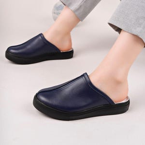 Peut inclure: Mules en cuir bleu marine avec une semelle noire. Les chaussures ont un bout arrondi et un profil bas. L'image montre les chaussures aux pieds d'une personne, suggérant un style de chaussures décontracté ou d'intérieur.