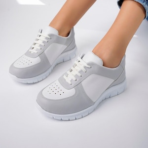 Può includere: Sneakers grigie e bianche con lacci bianchi. Le sneakers hanno una punta perforata e una suola bianca. Le scarpe sono su una superficie bianca. Le sneakers sono uno stile casual.
