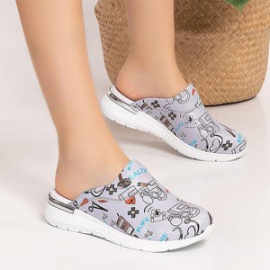 May include: Light gray clogs with a white sole and a medical-themed pattern. The clogs feature a variety of medical symbols and the words "#LIFE" and "HEALTH". The clogs are worn by a person sitting on a wooden stool.
