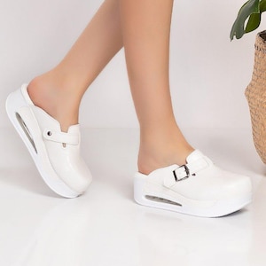 Könnte beinhalten: Weiße Clogs mit Schnallenverschluss und dicker Sohle. Die Clogs haben eine runde Spitze und ein kleines Luftloch. Das Bild zeigt auch einen schwarzen Jeansrock und einen geflochtenen Korb mit einer grünen Pflanze.