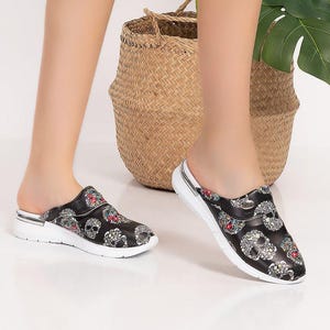 May include: Black slip-on clogs with a skull pattern and white soles. The clogs have a silver trim and are worn with a black skirt. A woven basket and a large green plant are in the background.