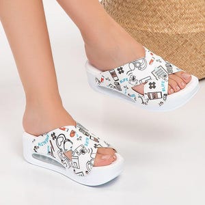 May include: White platform sandals with a graphic print featuring medical-themed illustrations and the word "LIFE". The sandals have a cross-strap design and a thick sole. The overall theme is healthcare and lifestyle.