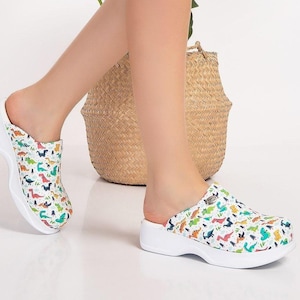 Peut inclure: Sabots blancs avec un motif de dinosaures colorés. Les chaussures à enfiler ont une semelle blanche épaisse et un motif représentant divers dinosaures verts, bleus, rouges et jaunes. Les sabots sont portés.