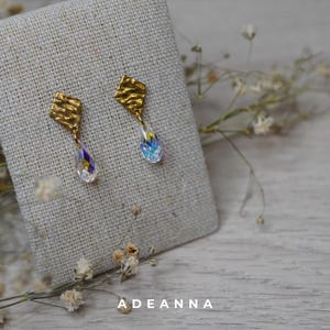 Könnte beinhalten: Ein Paar goldfarbene Ohrringe mit strukturiertem, quadratischem Oberteil und einem tropfenförmigen, irisierenden Kristallanhänger. Die Ohrringe werden auf einer beigen Leinenoberfläche präsentiert. Das Wort "ADEANNA" ist unten in Weiß gedruckt.
