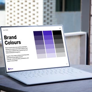 May include: A white laptop displaying a "Brand Colours" presentation with a color palette of purple, gray, and black. The laptop is open on a round, dark gray table outdoors, with a blurred background of a building.