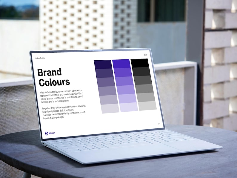 May include: A white laptop displaying a "Brand Colours" presentation with a color palette of purple, gray, and black. The laptop is open on a round, dark gray table outdoors, with a blurred background of a building.