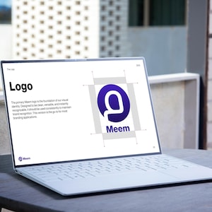May include: A white laptop displaying a logo presentation on a round, dark gray table. The screen shows the word "Logo" in bold black text, along with the Meem logo, a purple "M" inside a blue circle. The laptop is open, showcasing its keyboard.