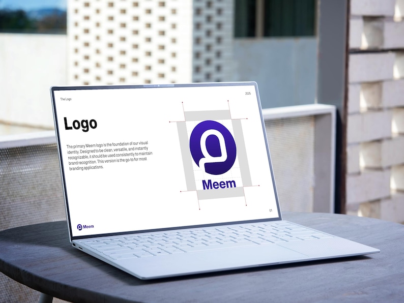 May include: A white laptop displaying a logo presentation on a round, dark gray table. The screen shows the word "Logo" in bold black text, along with the Meem logo, a purple "M" inside a blue circle. The laptop is open, showcasing its keyboard.