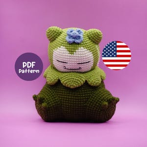 May include: A handmade green crochet plush toy with a white face and blue flower detail. The toy is sitting down with its eyes closed. The image includes a round graphic with the text "PDF Pattern" and an American flag.