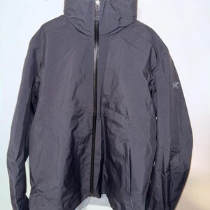 May include: A charcoal-grey jacket with a high, zipped collar. The jacket features a small logo on the left sleeve and a pocket on the right side. It is suspended from a wooden clothes airer against a white wall.