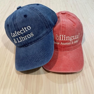 May include: Two baseball caps, one navy blue and one red. The navy cap has white text that reads "Cafecito & Libros." The red cap has white text that reads "bilingual Jane Austen & Gaby."