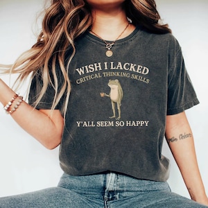 K&ouml;nnte beinhalten: Ein anthrazitfarbenes Kurzarm-T-Shirt mit einer Grafik eines stehenden Frosches, der eine kleine braune Tasse h&auml;lt. Der Text WISH I LACKED CRITICAL THINKING SKILLS Y'ALL SEEM SO HAPPY ist in hellbeiger Schrift &uuml;ber und unter dem Frosch abgebildet.