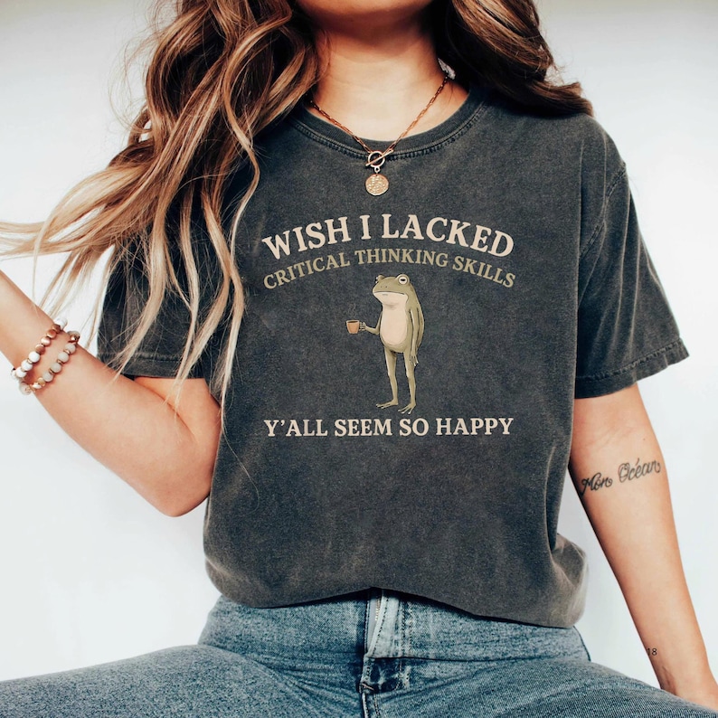 K&ouml;nnte beinhalten: Ein anthrazitfarbenes Kurzarm-T-Shirt mit einer Grafik eines stehenden Frosches, der eine kleine braune Tasse h&auml;lt. Der Text WISH I LACKED CRITICAL THINKING SKILLS Y'ALL SEEM SO HAPPY ist in hellbeiger Schrift &uuml;ber und unter dem Frosch abgebildet.