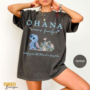 May include: Dark gray t-shirt with the word "OHANA" in light blue, featuring a cartoon character and the phrase "means family." Below, the text reads "nobody gets left behind or forgotten."