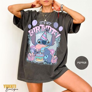 May include: Dark grey t-shirt with a cartoon character, birthday cake, and the text "IT'S MY BIRTHDAY". The design incorporates purple and pink balloons and gift boxes. The shirt is from Yukhti Boutique.